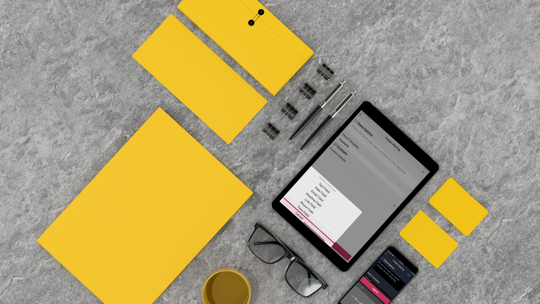 Office items organized into a grid pattern on a marbled, gray table. The items include two yellow business sized envelopes, a yellow paper, 4 binder clips, 2 pens, a tablet, eye glasses, a yellow mug with coffee, two yellow sticky notes, and a smartphone. The tablet and smartphone both show the WayAround app.