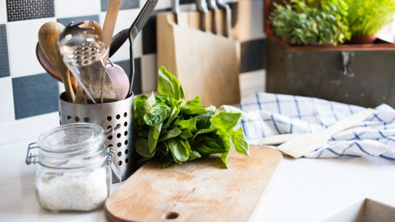 Closeup of a kitchen counter with a wooden cutting board, a metal container filled with kitchen utensils, and a clear glass container with salt. A bunch of fresh basil sits atop the cutting board. In the background are a couple of plants and a blue and white kitchen towel.