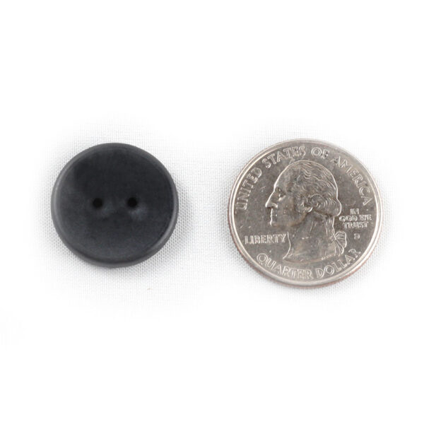A black two hole button next to a quarter. The button i s slightly smaller than the quarter.