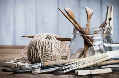 A wooden surface holds various hobby implements, tan yarn with clothespins and various wooden knitting tools. At the forefront is a knocked over mason jar of paint brushes with wooden handles.