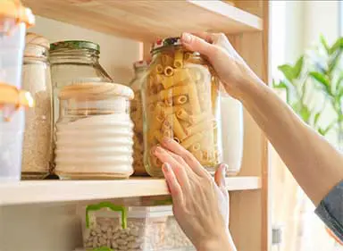 A person reaches up to pull a large mason jar of dry pasta from a shelf also containing other dry goods.