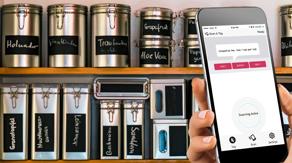 Photograph illustrating how WayAround can help people with vision disorders to distinguish between metal canisters in the kitchen. Someone is holding a phone in front of a shelf with various canisters. The WayAround app is on the screen with text that reads Grapefruit tea. Use 1 tsp per cup